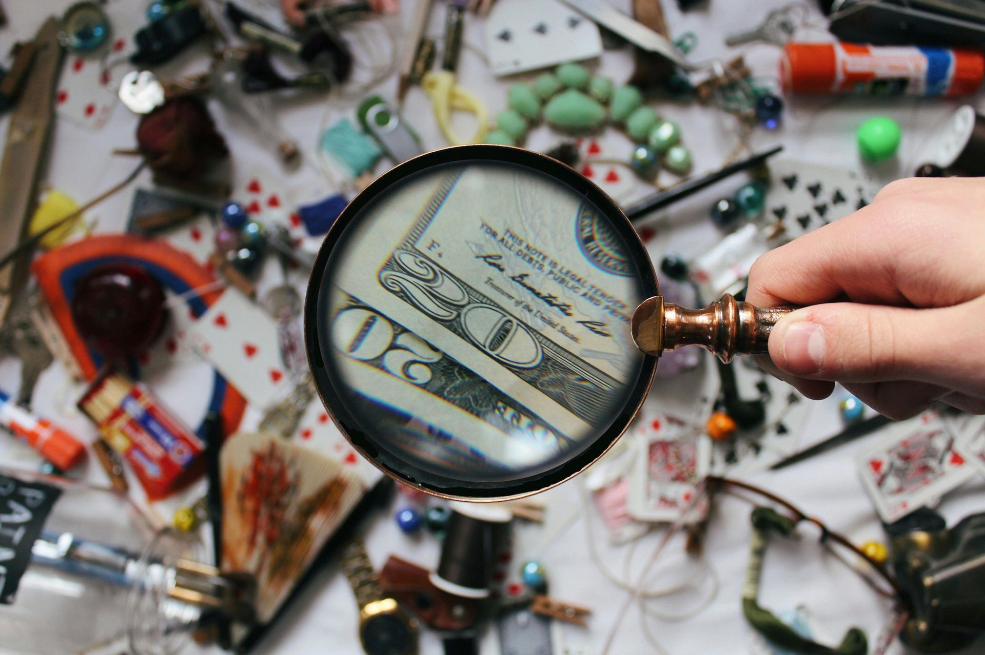 Hand holding magnifying glass over a twenty-dollar bill, amidst a clutter of various small objects.