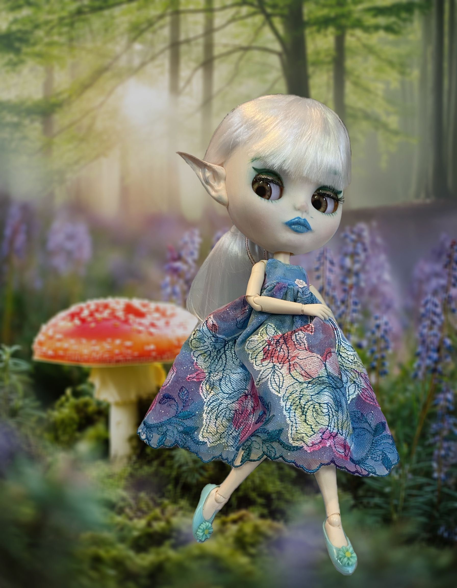 Pop met zilverkleurig haar en puntige oren in een blauwe jurk met patroon, geposeerd in een bosscène met paddenstoelen.