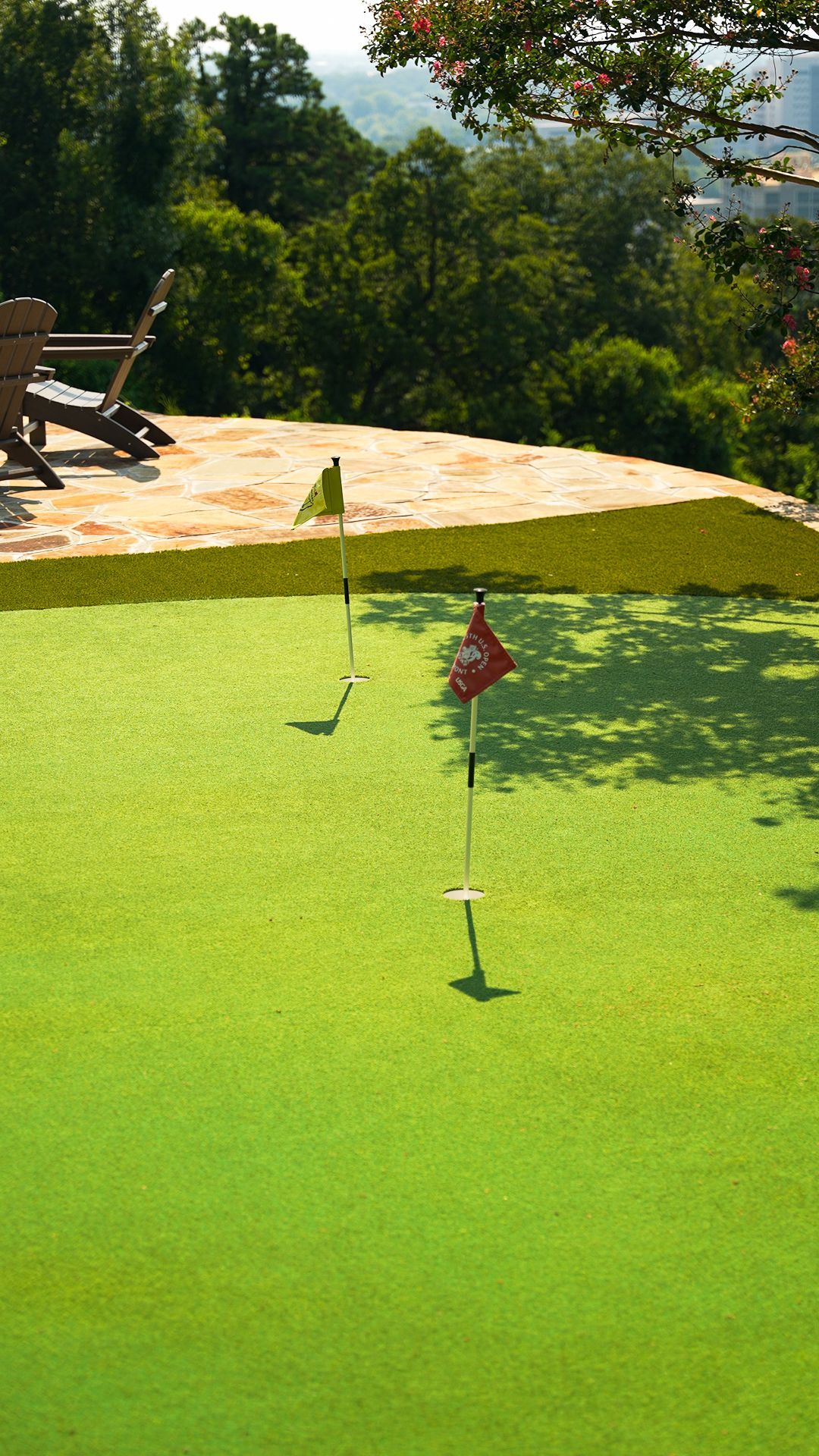 Lawn putting green with a flagstick beside a patio and trees in the background