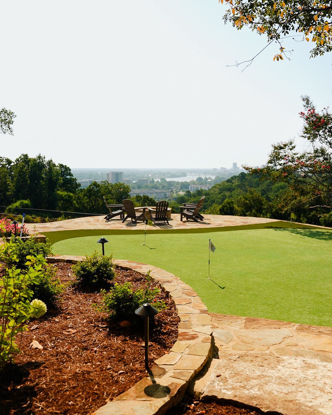 Park overlook with green lawn, stone path, benches, and distant city view under a bright sky