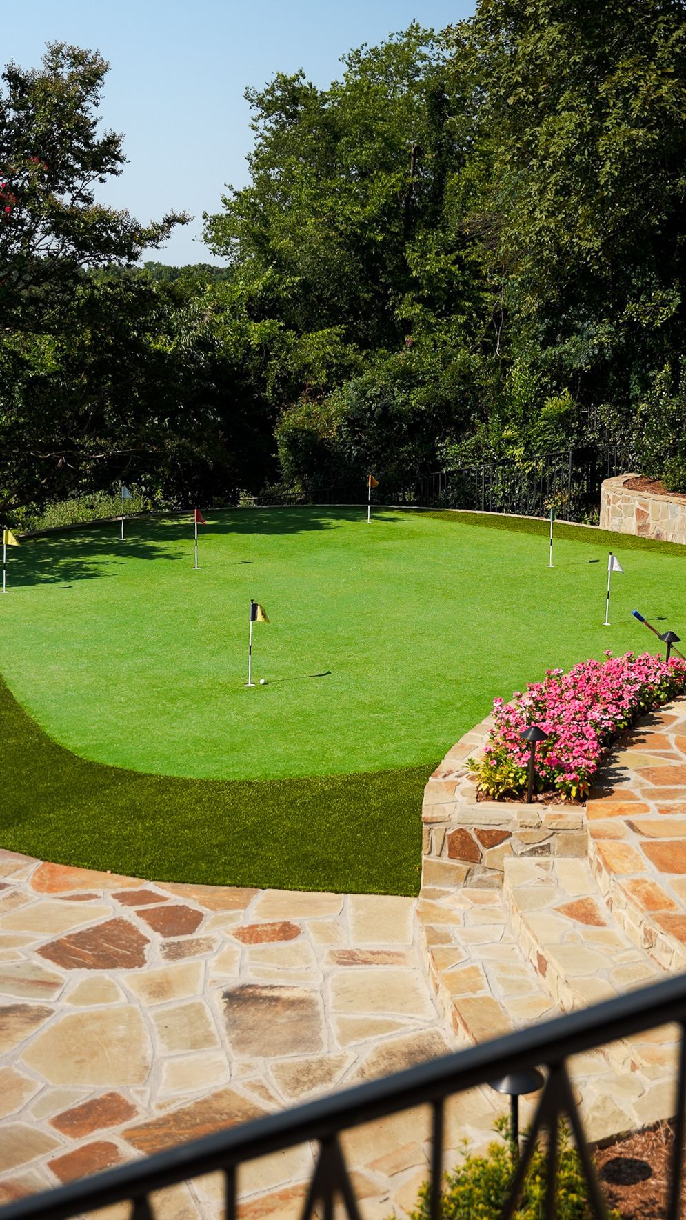 Sunny lawn with two sprinkler heads, stone patio, trees, and pink flowers bordering the garden