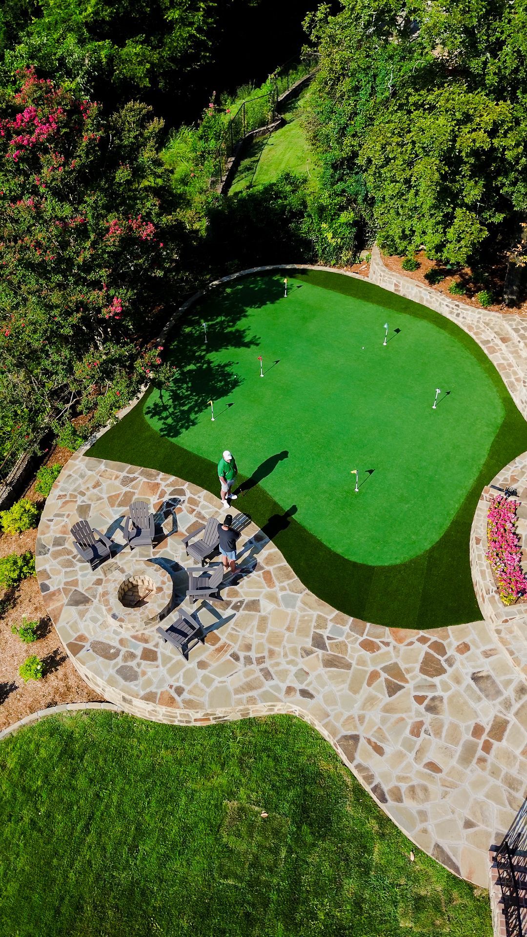 Aerial view of a mini golf green with a rock feature and stone walkway surrounded by lush greenery.