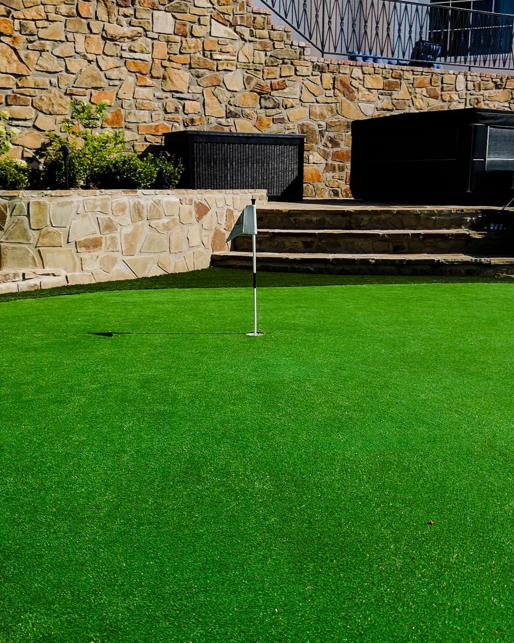 Green putting turf with a golf hole flag beside stone steps and wall outdoors