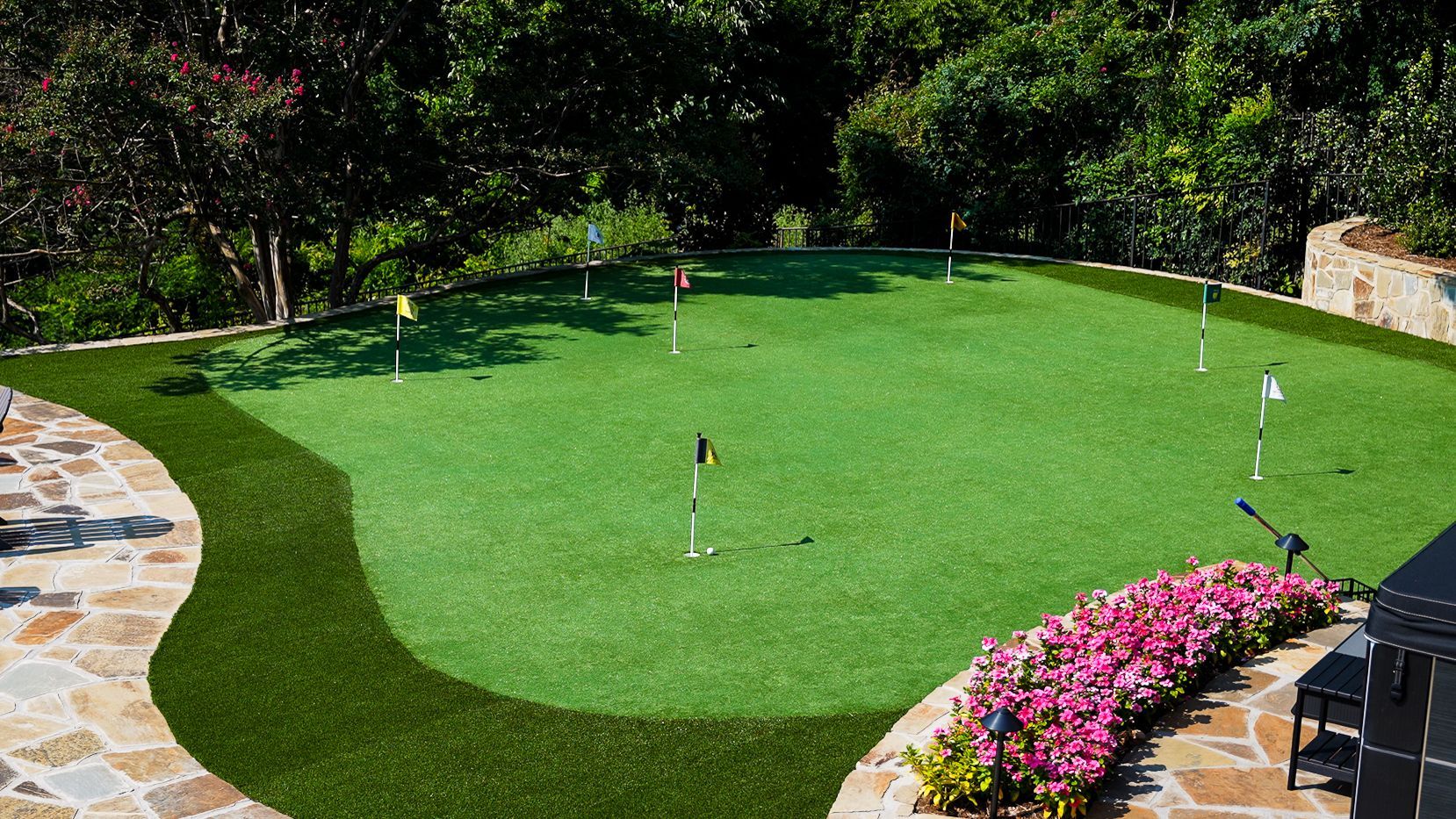 Backyard putting green with golf flags, surrounded by stone paths and pink flowers
