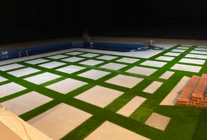 An outdoor event venue with rows of white square tables on green turf, lit at night.