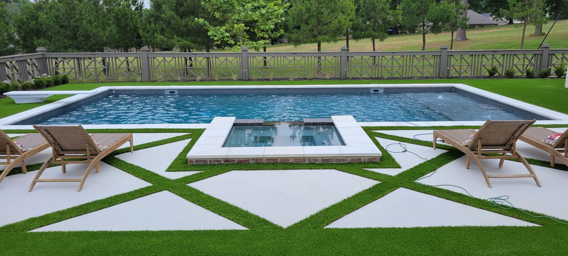 Modern backyard pool with geometric stone patio, two lounge chairs, and greenery surrounding the water.