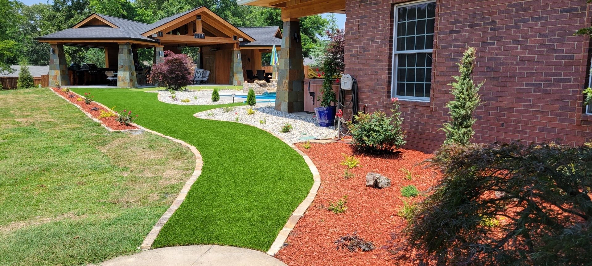 Landscaped backyard with a curving green lawn, red mulch beds, and a wooden gazebo by a brick house