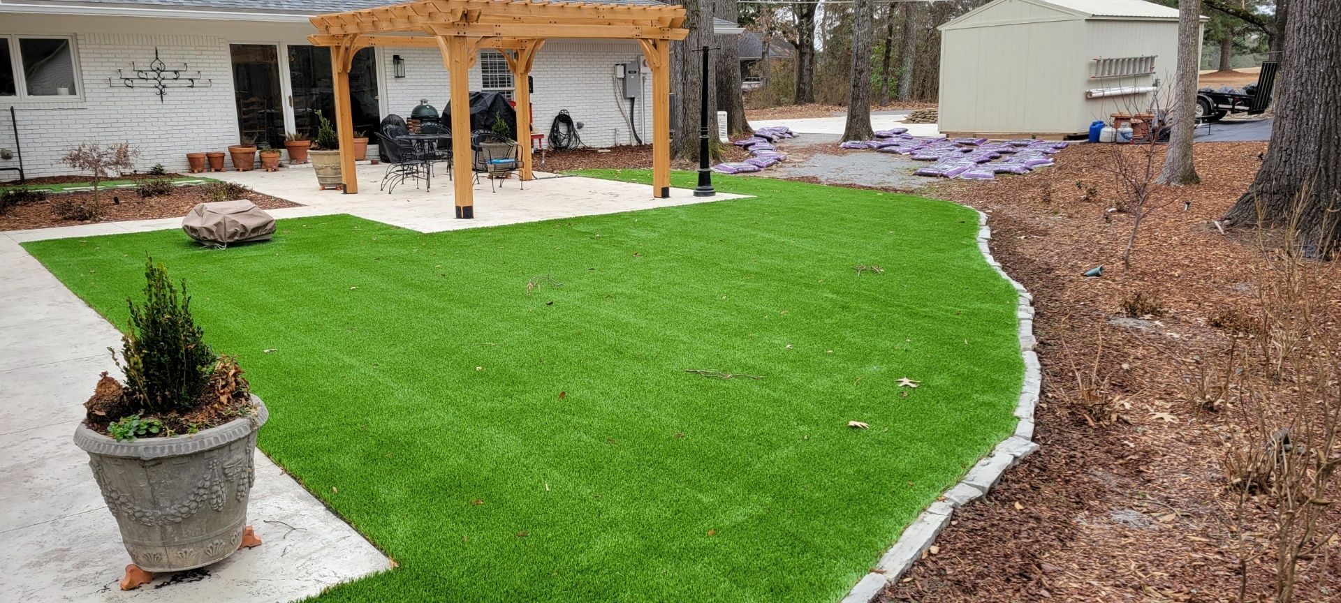 Landscaped yard with bright green lawn, patio pergola, and large trees along the edge