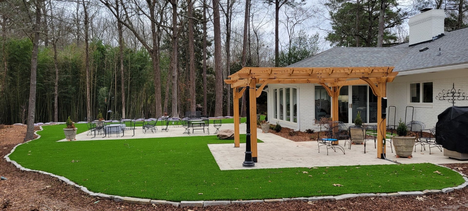 Backyard patio with a wooden pergola, green lawn, paver edging, and wooded trees beyond the house