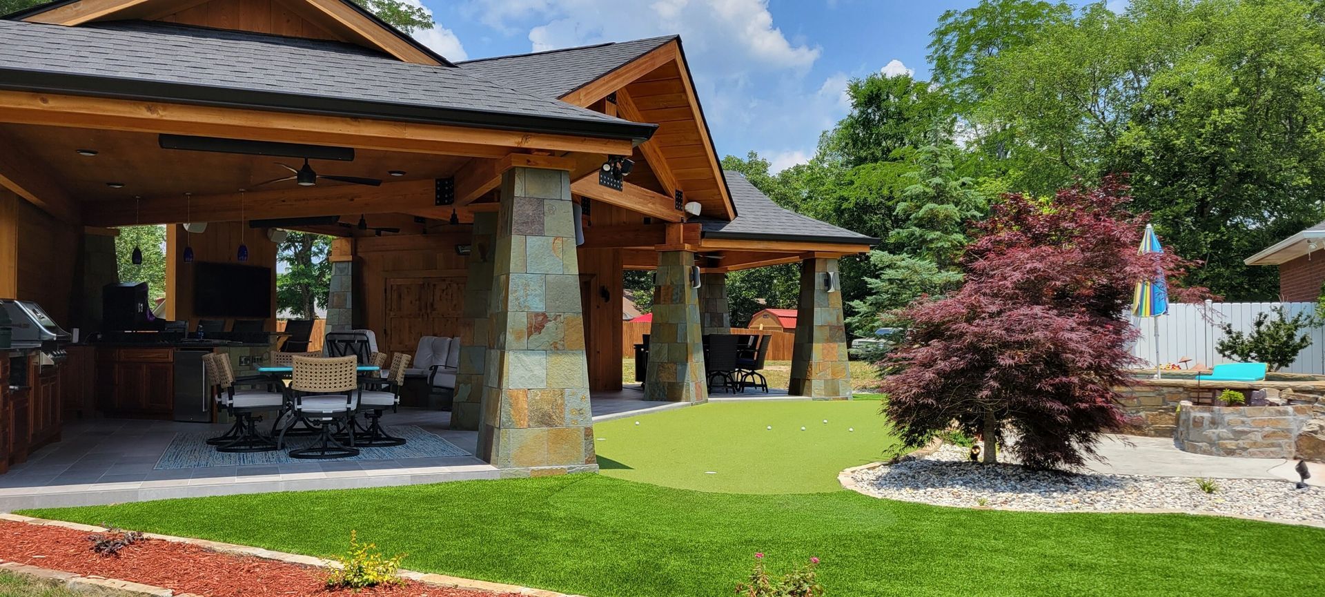 Covered patio and landscaped yard with stone columns, wooden beams, green lawn, and a red shrub