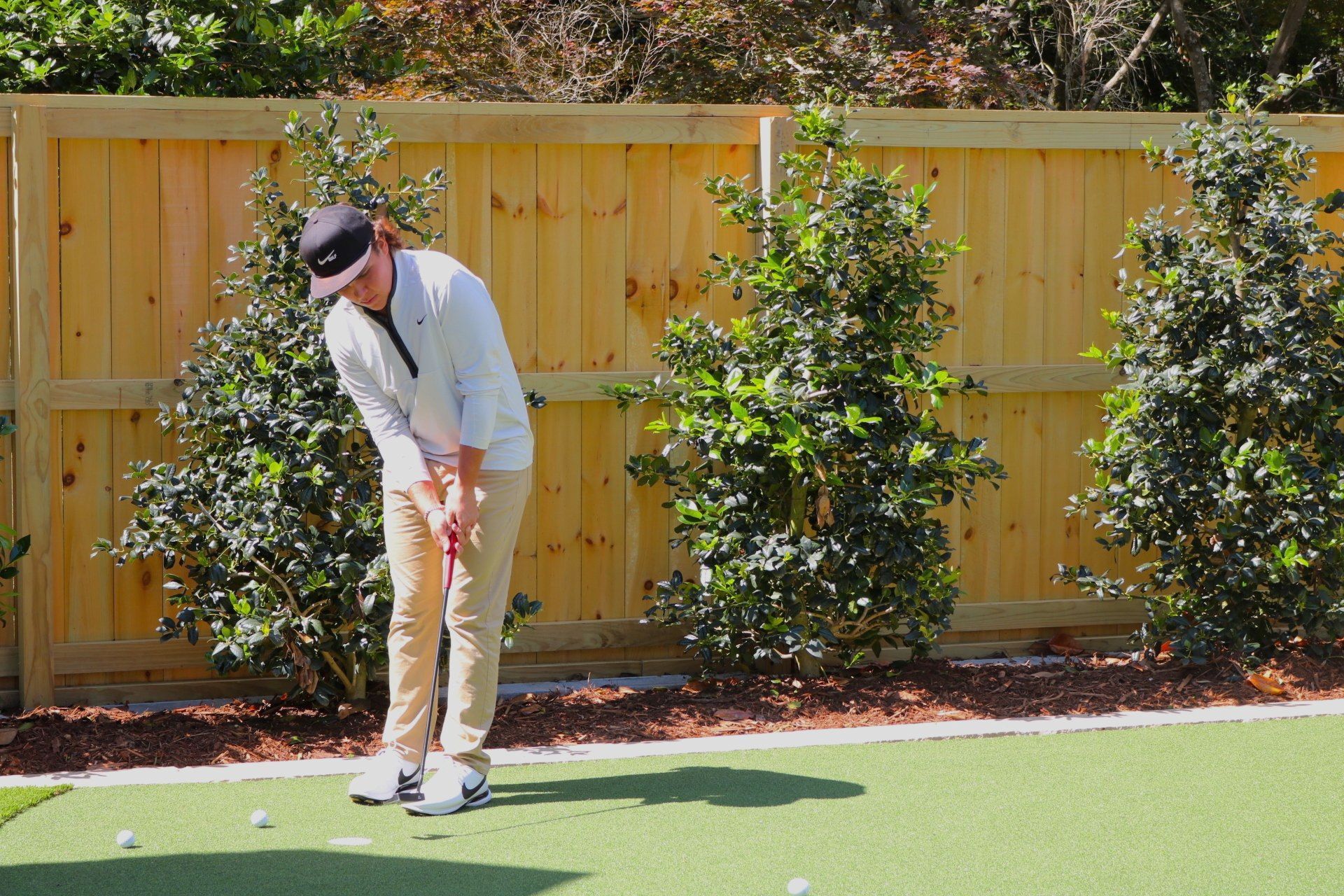 A man is swinging a golf club on a golf course in front of a wooden fence.