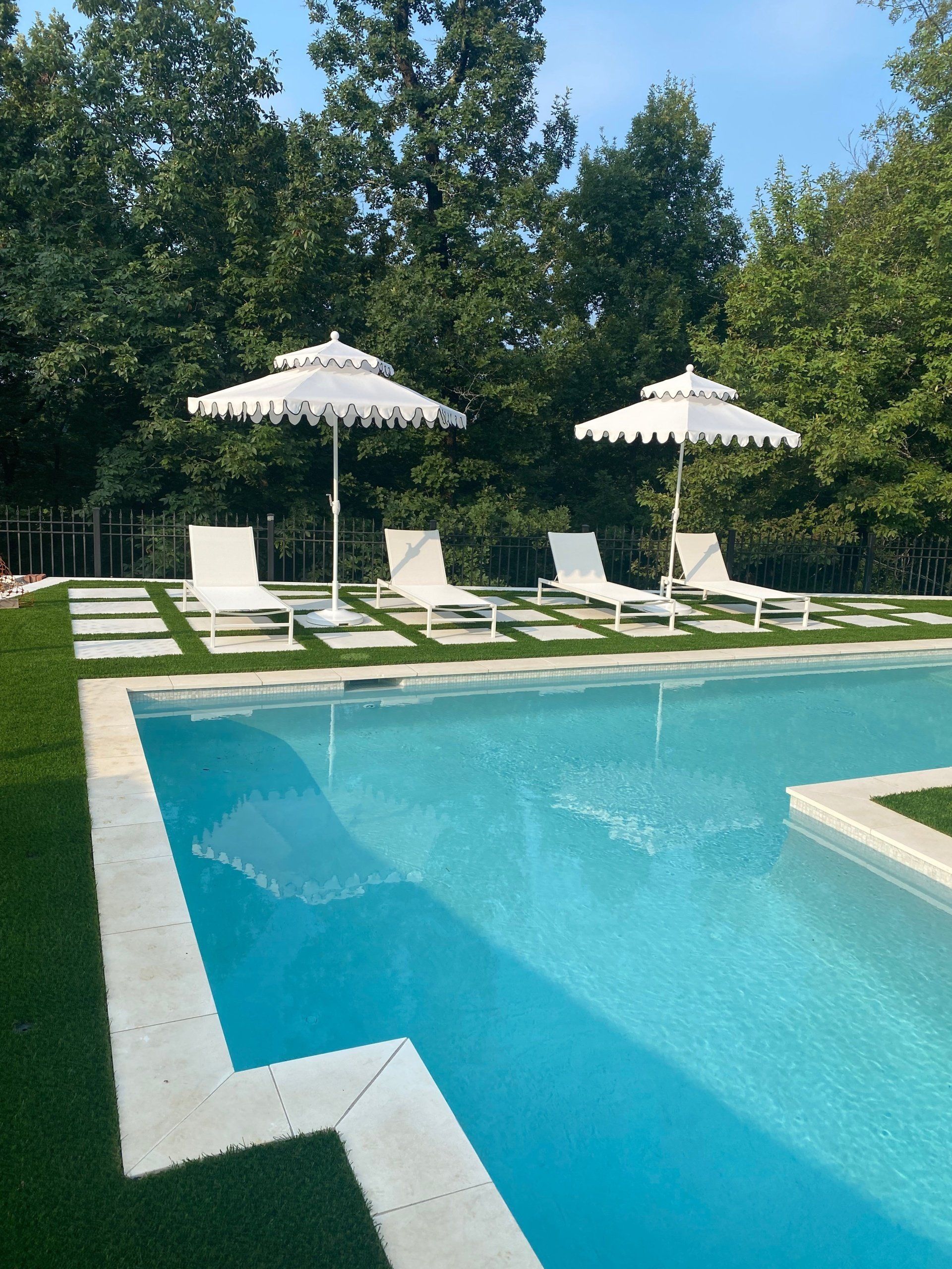 Poolside patio with white lounge chairs, parasols, and trees beside a turquoise swimming pool