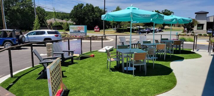 Outdoor café seating with teal umbrellas and artificial grass beside a sidewalk and parked cars