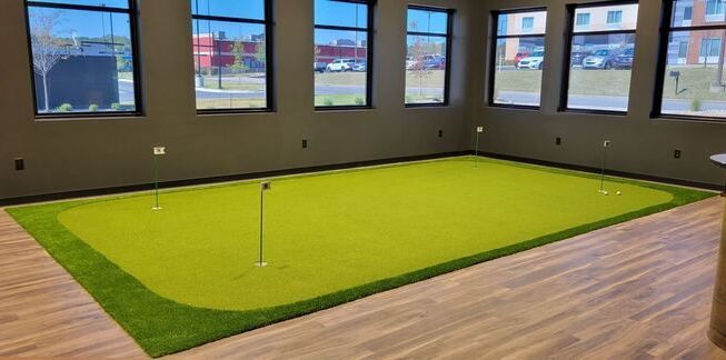 Indoor putting green with bright green turf in a room with large windows and wood flooring