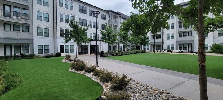Modern apartment courtyard with white buildings, green lawn, trees, and a paved walkway