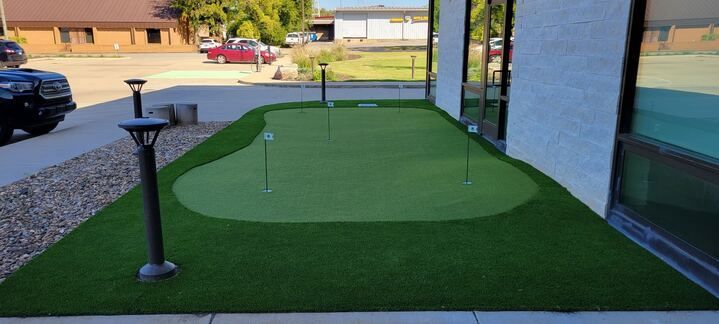 Outdoor putting green beside a building, with two black path lights and a gravel parking area nearby