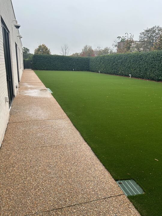 Wet patio beside a white house, overlooking a green lawn and tall hedge under cloudy skies