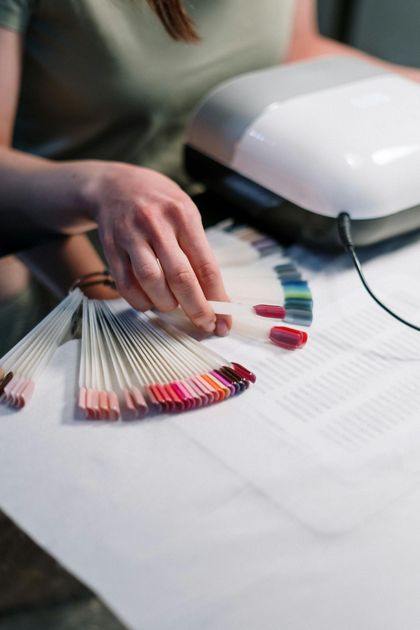 Persona eligiendo un color de esmalte de uñas de un expositor con forma de abanico, cerca de una lámpara de curado.