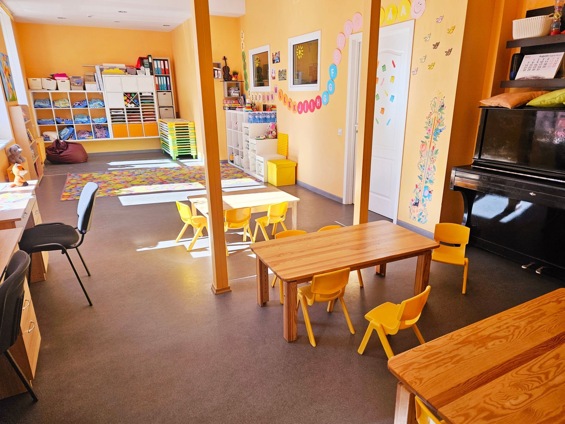 Bērnu aprūpes klase ar galdiem, krēsliem, rotaļlietām un klavierēm gaišā telpā
