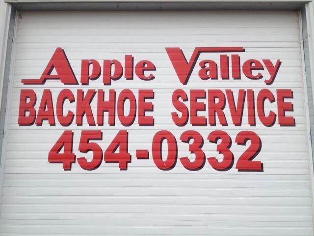 Red sign reading “Apple Valley Backhoe Service 454-0332” on a white metal wall