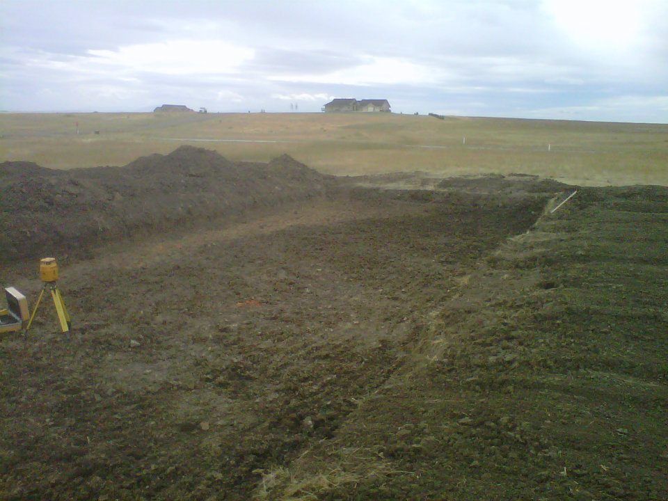 A muddy rural field with a dirt path leading toward distant farm buildings under a cloudy sky