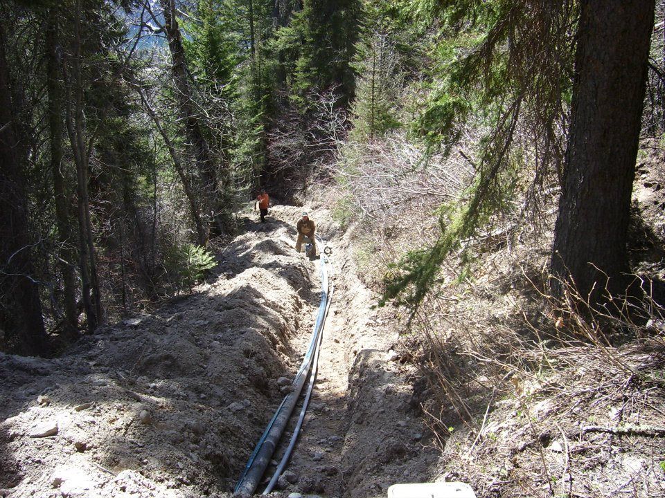 Two workers in a forest trench laying a blue pipeline through muddy ground