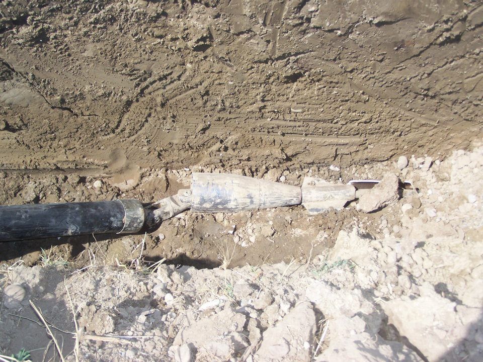Damaged underground pipe with black hose and white insulation exposed in a dirt trench