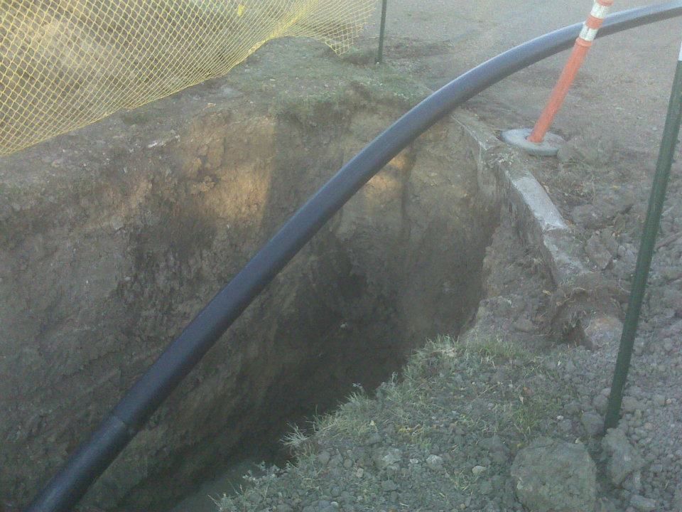 Black pipe runs through a dug trench beside a construction barrier and orange stake.