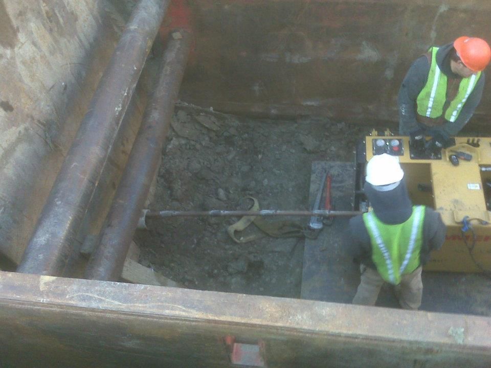 Workers in safety vests inspect a deep excavation site with pipes and equipment.