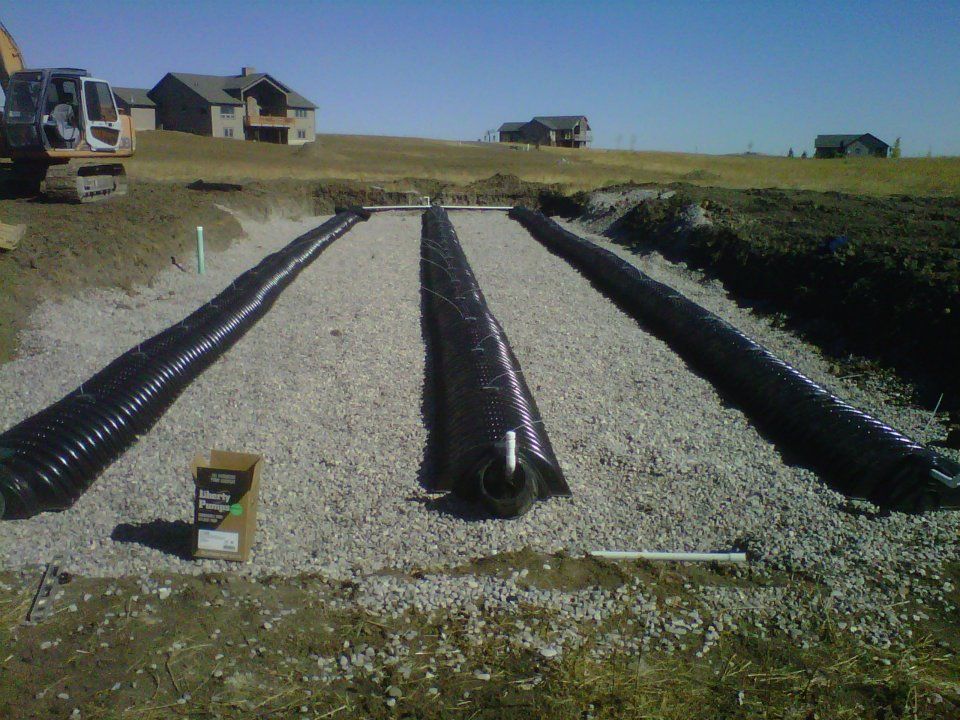Gravel drainage trenches lined with black plastic in a grassy field with houses in the background