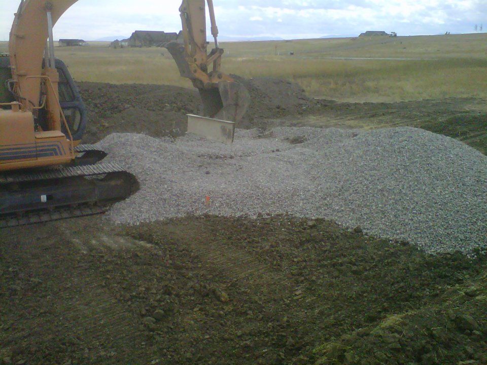 Excavator dumping gray gravel onto a pile in an open grassy field