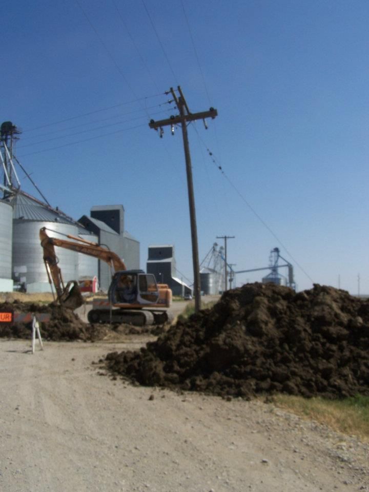 Excavator and dirt piles along a dusty construction site with utility poles and industrial silos in the background