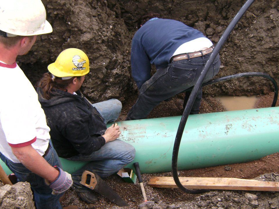 Workers in hard hats repair a large green underground pipe in a dirt trench.