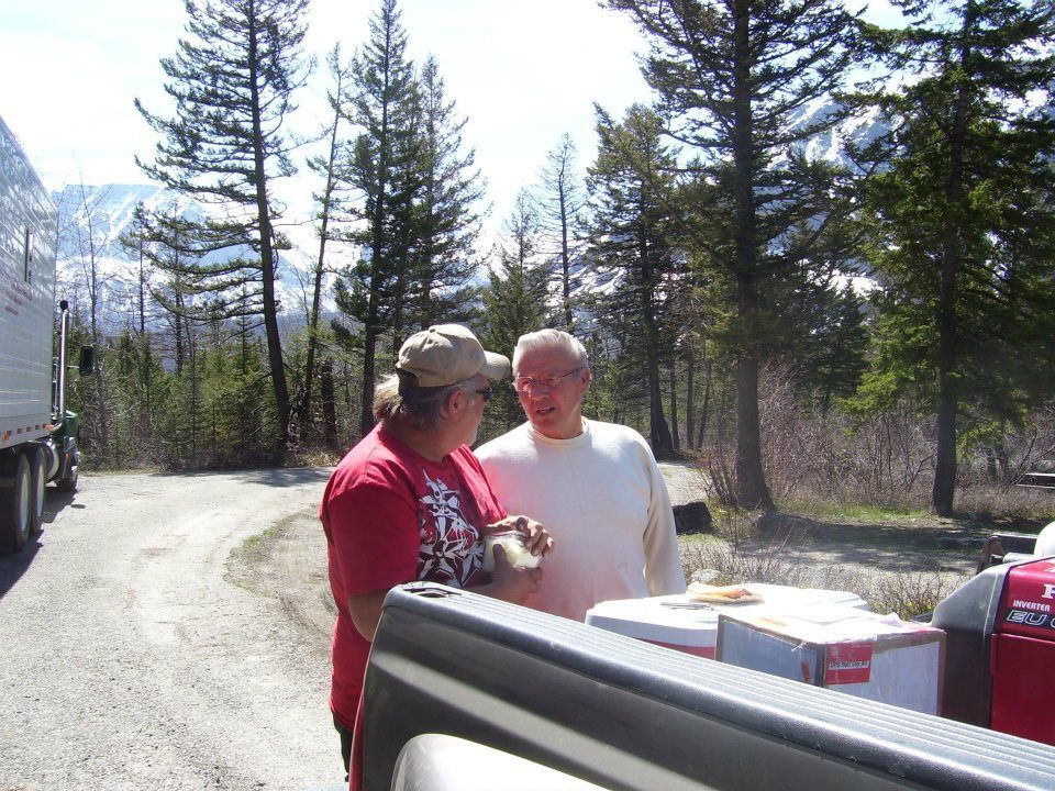 Two people talking beside a truck at a wooded campsite with mountains in the background.