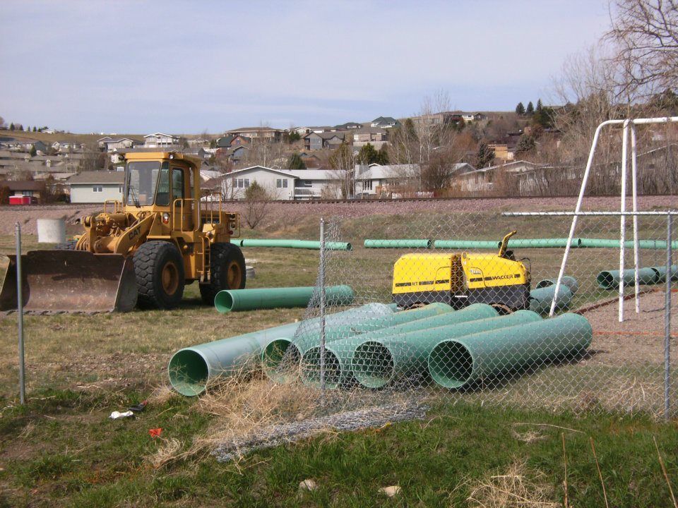 Construction equipment and large green pipes in a fenced field, with houses in the background.