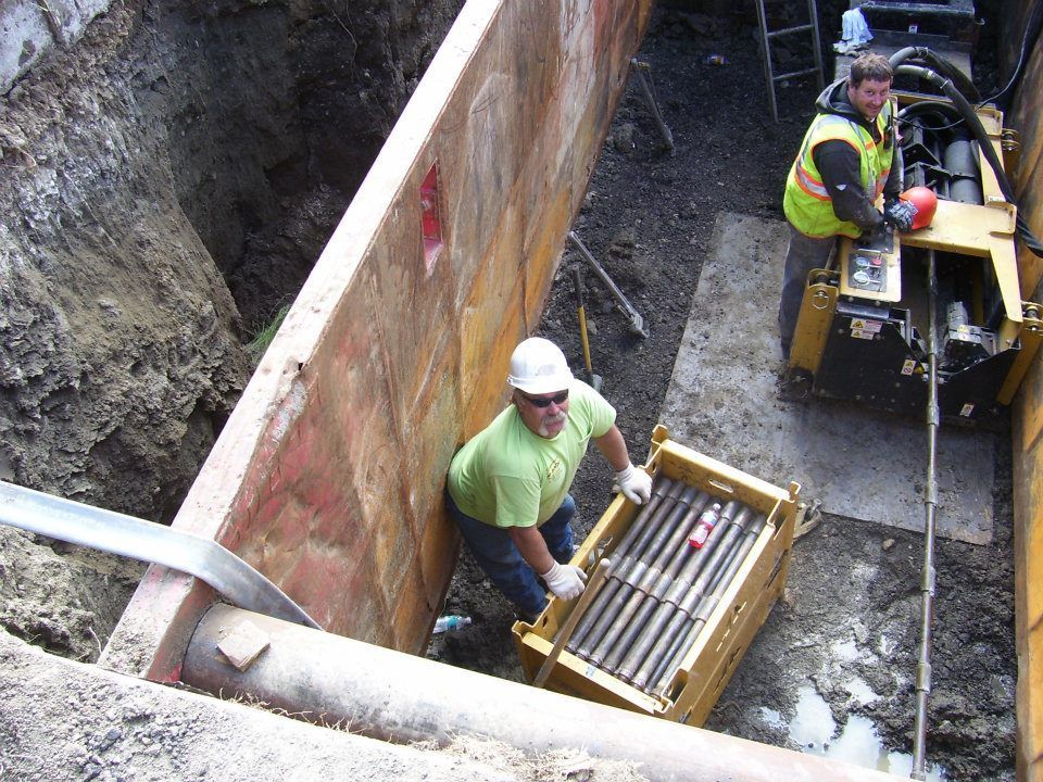 Workers using construction equipment in a trench beside a rusted retaining wall