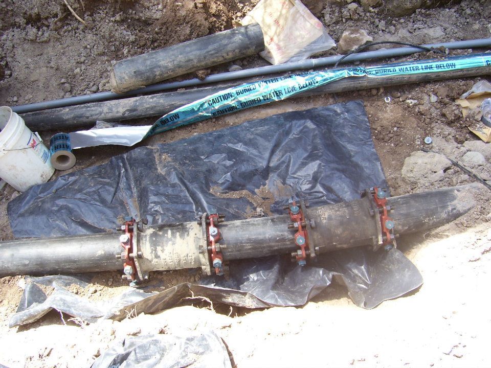 Damaged underground pipe section with couplings, wrapped in black plastic on dirt ground