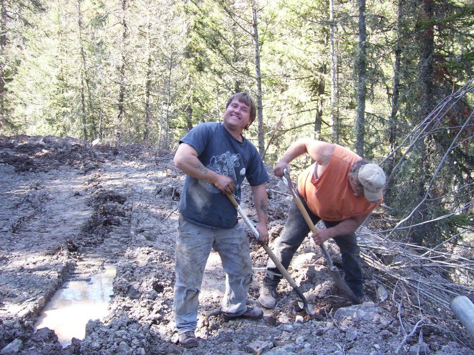 Two people working outdoors in a wooded area, shoveling muddy ground near a forest trail.