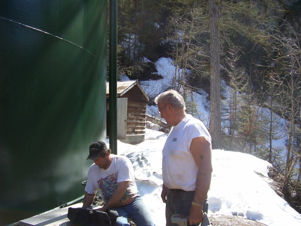 Two men outdoors by a green wall and snowy hillside, one seated and one standing nearby.