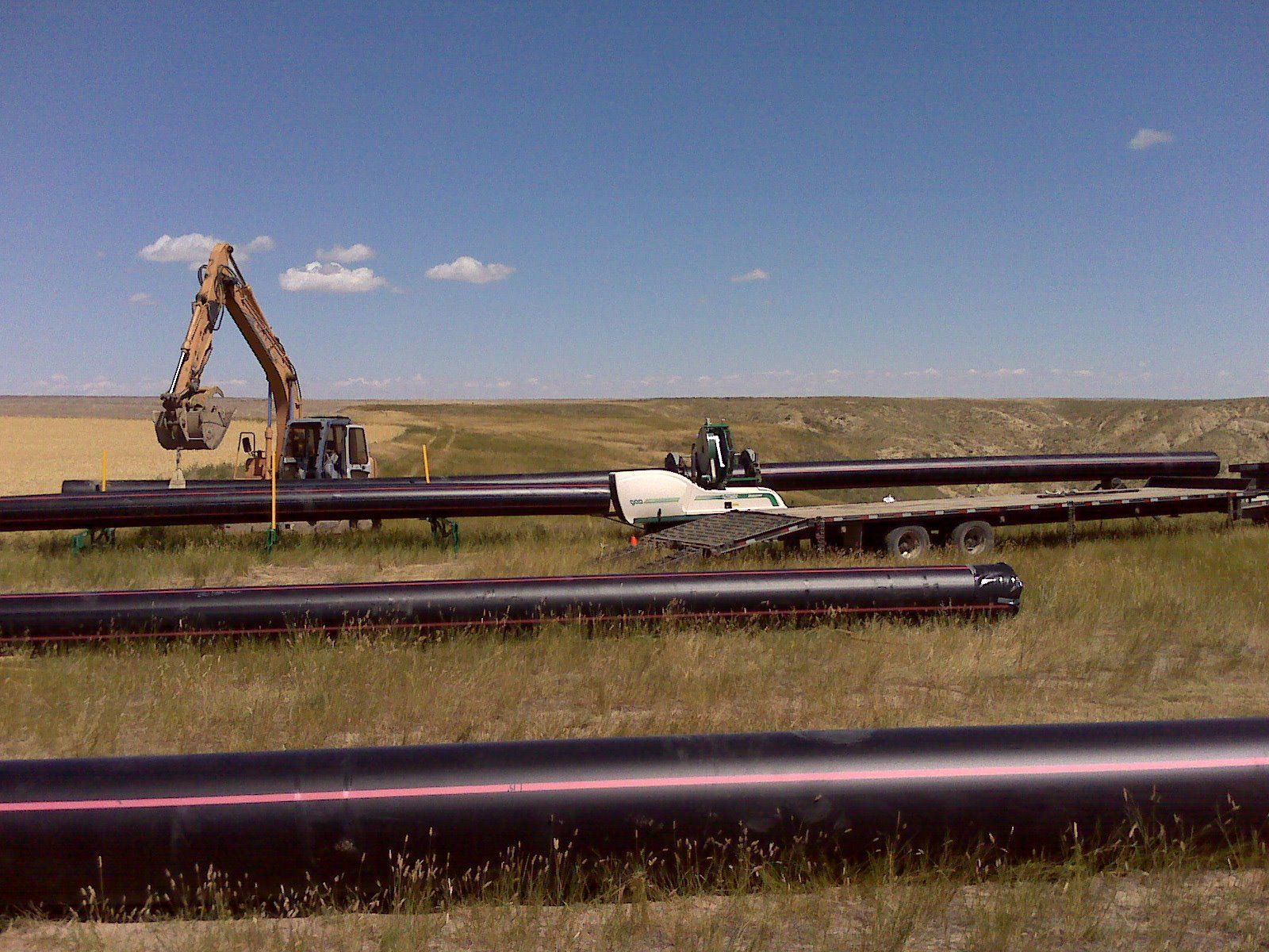 Large black pipelines in a grassy field with an excavator and work truck under a blue sky
