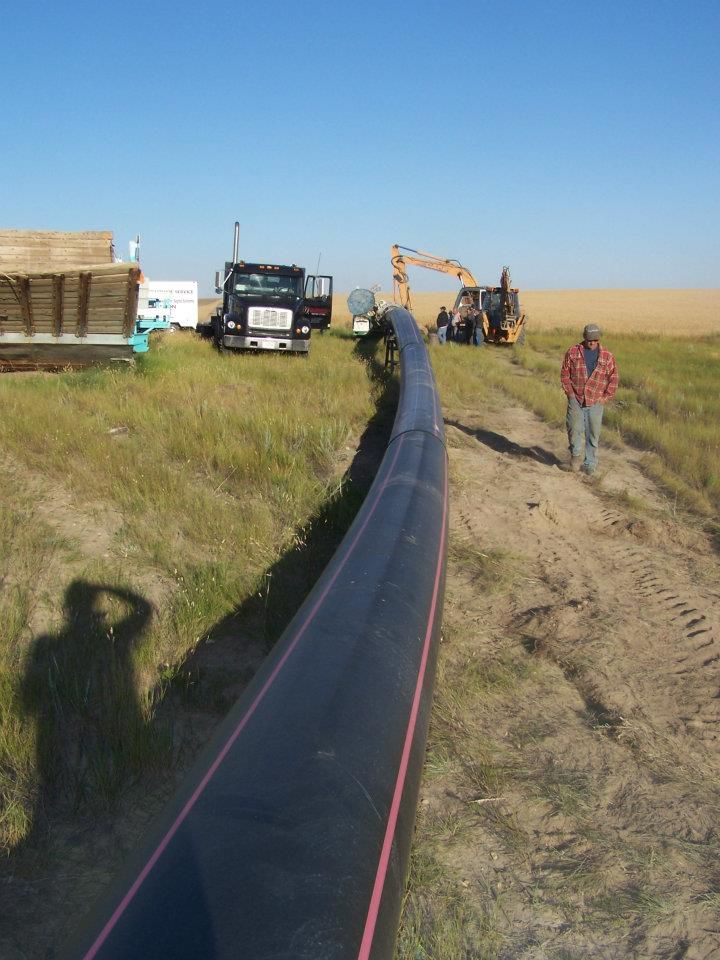 Workers laying a black pipeline across a grassy dirt field with trucks and an excavator nearby