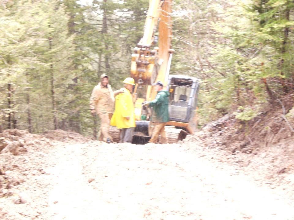 Construction crew with backhoe on a muddy forest road, one worker in yellow vest standing beside the machine