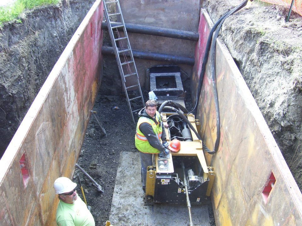 Workers and machinery in a narrow excavation trench with a ladder and concrete walls