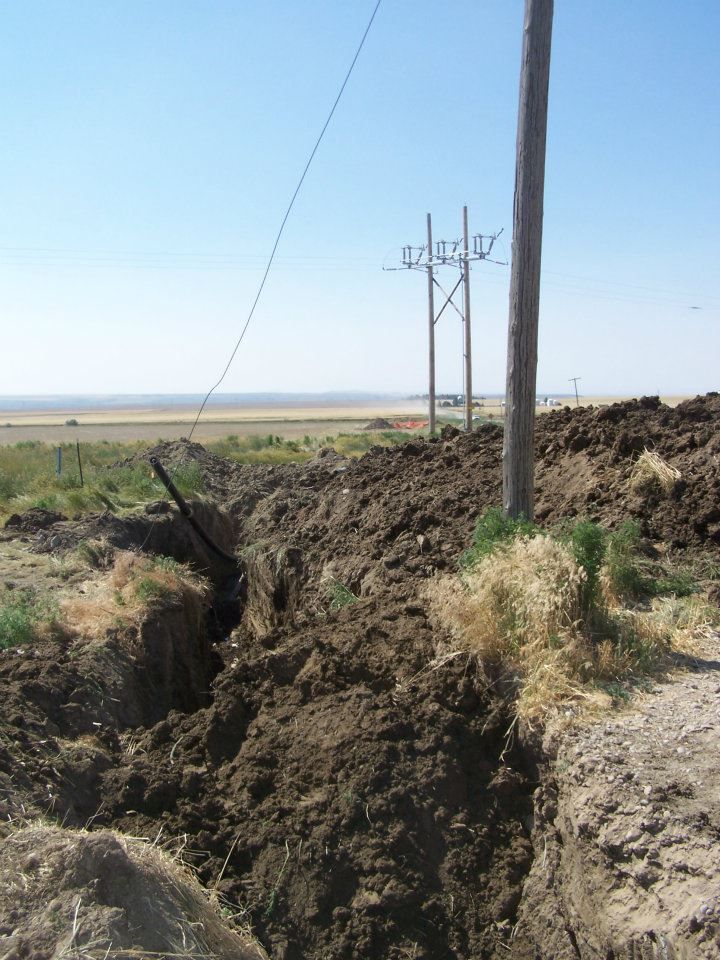 Deep trench cut through a grassy field beside utility poles under a clear blue sky