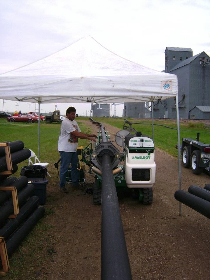 Person operating a machine under a white canopy tent beside long black pipes outdoors