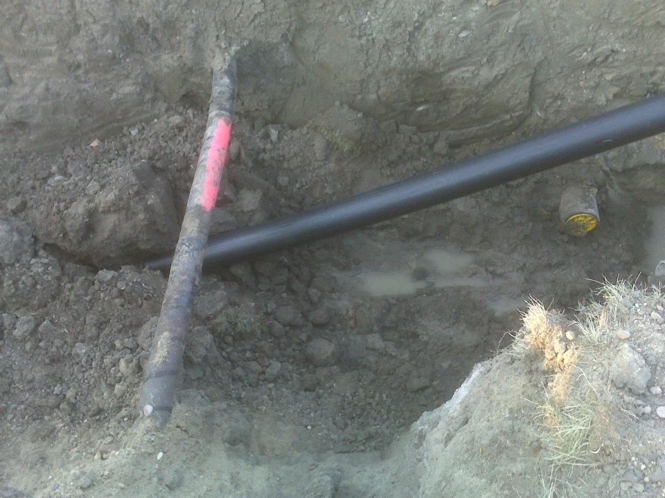 Black pipe and pink-marked tool handle crossing in a dirt excavation trench