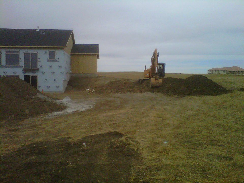 Construction site beside a house, with an excavator and dirt piles on a cloudy day.