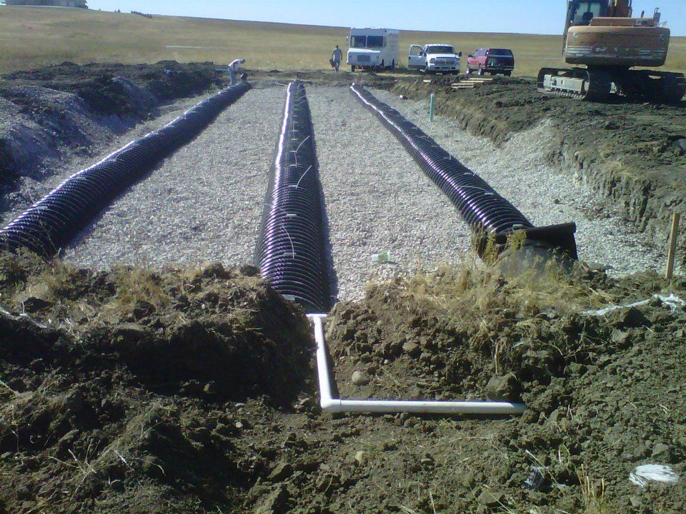 Trenched field with gravel drainage pipes laid in parallel under construction, with trucks and excavator nearby