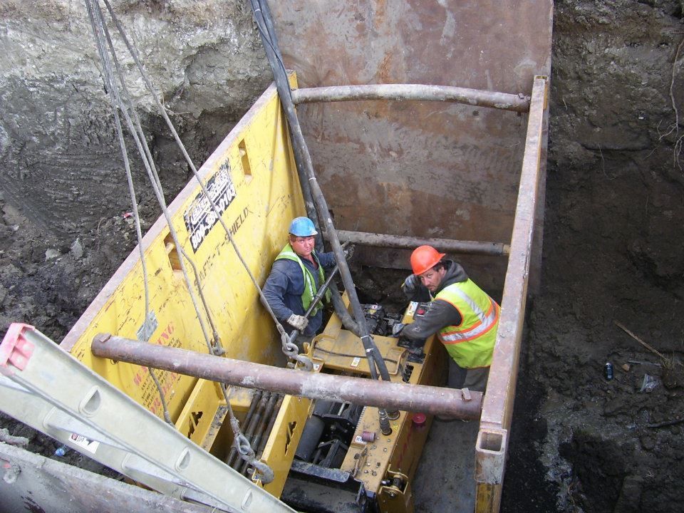 Two workers in hard hats operate equipment in a deep excavation with yellow shoring panels.