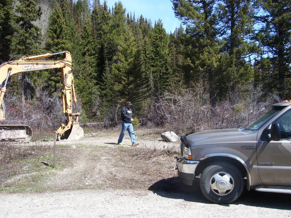 Excavator and person near a parked pickup truck by a wooded roadside area